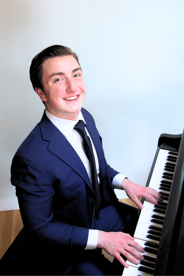 Samuel Hedgpeth smiling while seated at a piano in a blue suit.
